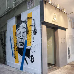 Photo of a building corner whose wall bears a mural of a man in glasses, with yellow and blue stripes and a bitcoin symbol.