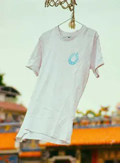 Photo of a white T-shirt with a small blue Serum logo, hanging outdoors in front of a temple roof.