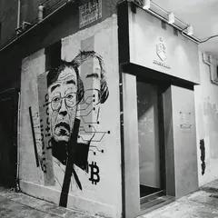 Animated black-and-white photo of a building corner whose wall bears a mural of a man in glasses and a bitcoin symbol.